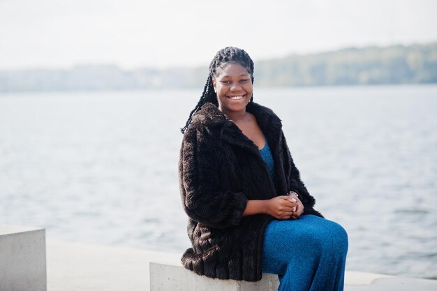 Afro-Amerikaanse donkere huid plus size model poseerde in een blauwe glanzende jurk en zwarte bontjas zittend op een stenen kubus tegen de zeezijde