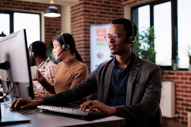 Afro-Amerikaanse hulplijnmedewerker die werkt bij de receptie van het callcenter met meerdere monitoren. Mannelijke operator die telecommunicatie gebruikt om klanten te helpen bij klantenservice, extern netwerk.