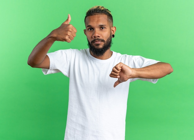 Afro-Amerikaanse jonge man in wit t-shirt kijken camera verward duimen opdagen