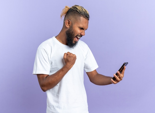 Afro-Amerikaanse jonge man in wit t-shirt met smartphone balde vuist gelukkig