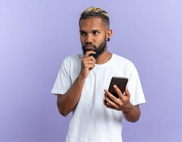 Afro-Amerikaanse jonge man in wit t-shirt met smartphone opzij kijkend verbaasd over blauwe achtergrond