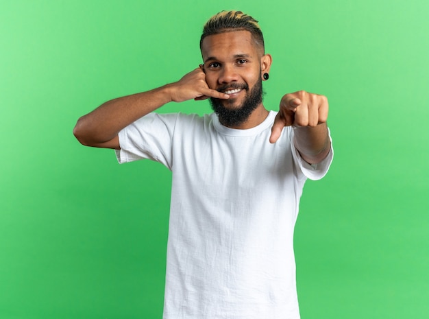 Afro-Amerikaanse jonge man in wit t-shirt wijzend met wijsvinger naar camera maken bel me gebaar glimlachend vriendelijk permanent over groene achtergrond