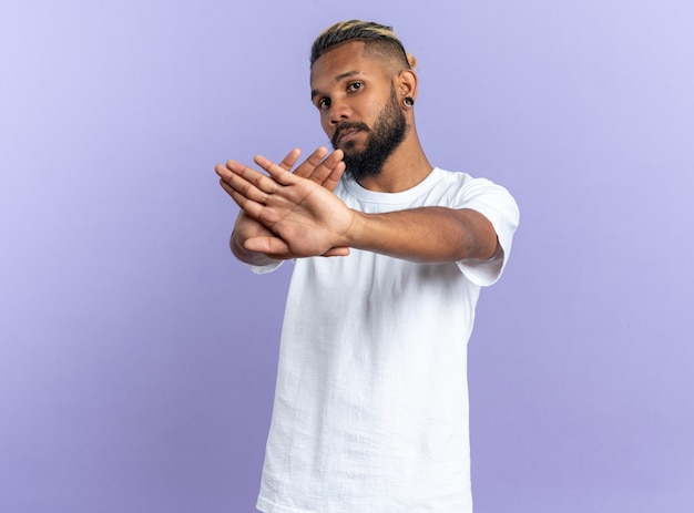 Afro-Amerikaanse jongeman in wit t-shirt die naar camera kijkt met een serieus gezicht dat een stopgebaar maakt met handen die over een blauwe achtergrond staan