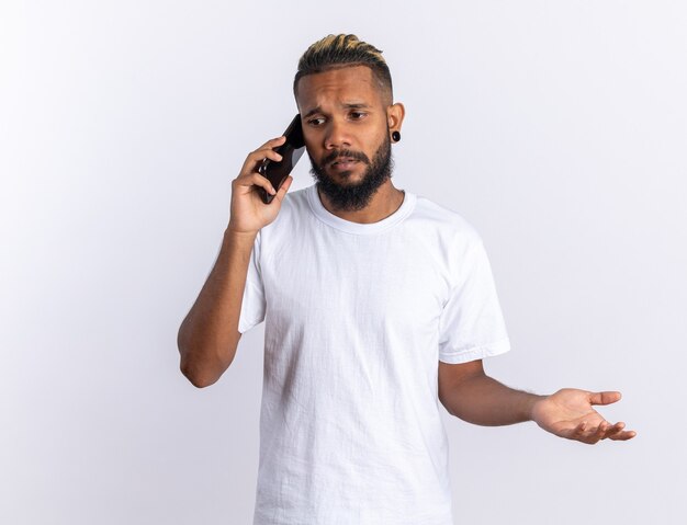 Afro-Amerikaanse jongeman in wit t-shirt die verward kijkt terwijl hij op een mobiele telefoon praat die over een witte achtergrond staat