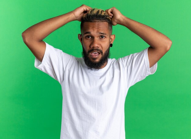 Afro-Amerikaanse jongeman in wit t-shirt kijkend naar camera die zijn haar trekt en gefrustreerd raakt