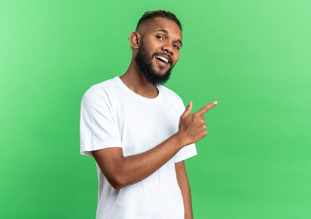 Afro-Amerikaanse jongeman in wit t-shirt kijkend naar camera glimlachend vrolijk wijzend met wijsvinger naar de zijkant