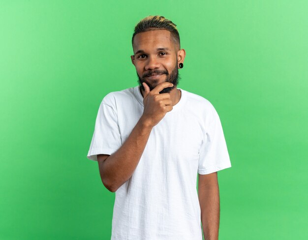 Afro-Amerikaanse jongeman in wit t-shirt kijkend naar camera met hand op zijn kin glimlachend vrolijk staande over groene achtergrond