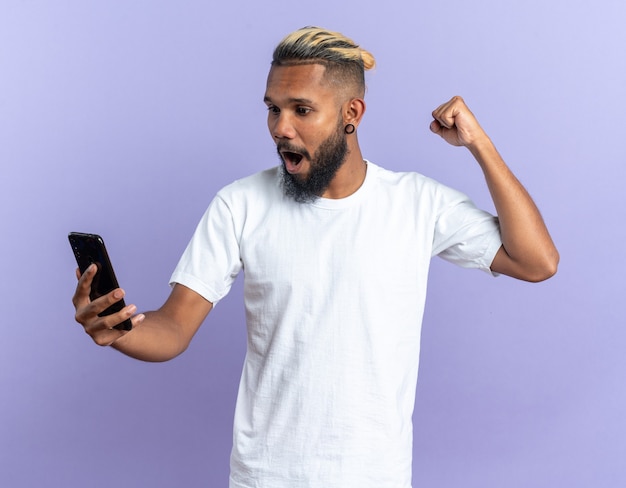 Afro-Amerikaanse jongeman in wit t-shirt met smartphone gebalde vuist blij en opgewonden schreeuwend blij met zijn succes staande over blauwe achtergrond