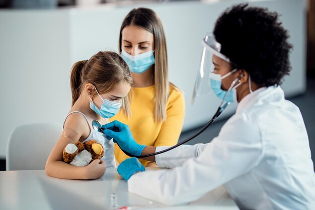 Afro-Amerikaanse kinderarts die een klein meisje onderzoekt met een stethoscoop tijdens een pandemie van het coronavirus