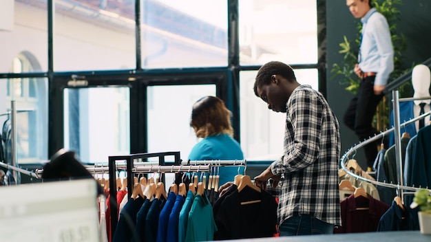 Gratis foto afro-amerikaanse klant winkelt voor een zwart shirt en analyseert modieus kledingmateriaal in een moderne boetiek. shopaholic man koopt vrijetijdskleding, kijkt naar hangers met nieuwe modecollectie