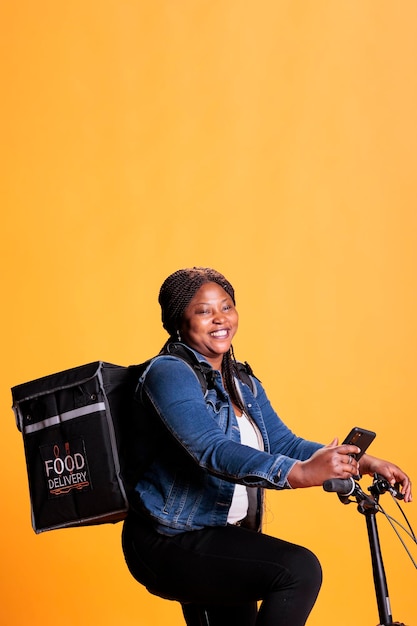 Afro-amerikaanse koerier die op de fiets rijdt terwijl hij afhaalmaaltijden aflevert aan de klant, adres controleert op de afhaalapp van de telefoon, staande op gele achtergrond in de studio. afhaalservice en concept