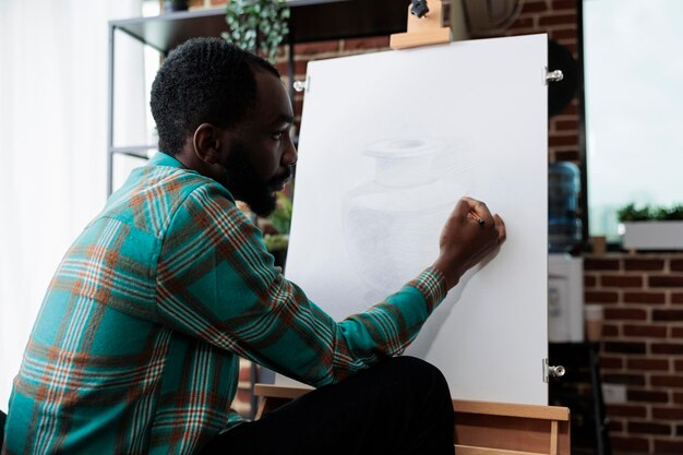 Afro-Amerikaanse kunstenaar vaasmodel tekenen op wit canvas werken bij illustratie techniek met behulp van grafisch potlood tijdens kunstles in creativiteit studio. Man schetst masterprice die nieuwe vaardigheid ontwikkelt
