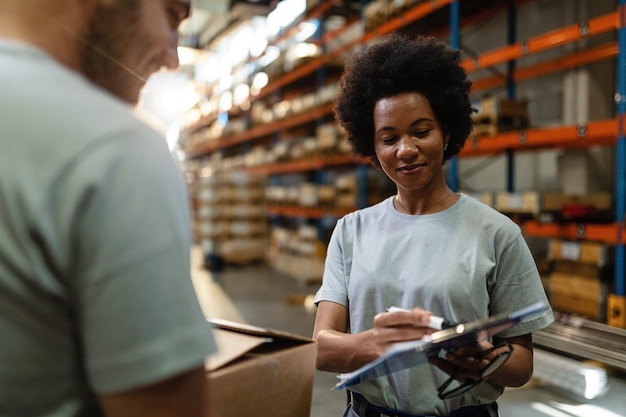 Afro-Amerikaanse magazijnmedewerker die aan haar collega een checklist met producten voor de verzending laat zien terwijl ze in het distributiemagazijn werkt