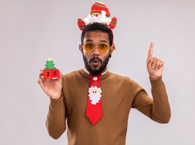 Afro-Amerikaanse man in bruine trui en kerstman rand op hoofd met grappige rode stropdas speelgoed blokjes met datum vijfentwintig kijken camera verrast tonen wijsvinger staande op witte achtergrond