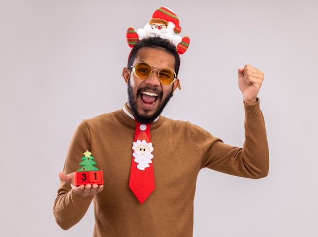 Afro-amerikaanse man in bruine trui en santa rand op hoofd met grappige rode stropdas speelgoed blokjes met nieuwjaar datum kijken camera blij en opgewonden gebalde vuisten permanent op witte achtergrond