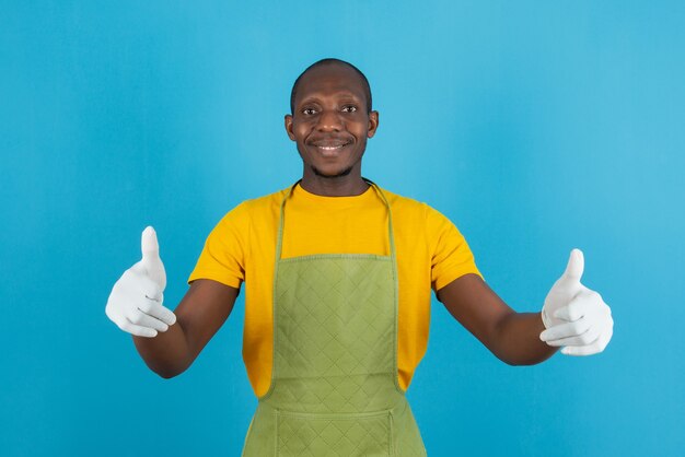 Afro-Amerikaanse man in groene schort met duimen op blauwe muur