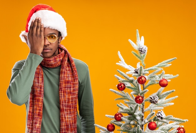 Afro-Amerikaanse man in kerstmuts en sjaal om nek camera kijken met ernstig gezicht voor één oog met een hand die naast een kerstboom staat op oranje achtergrond