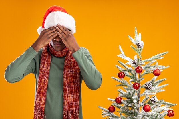 Afro-Amerikaanse man in kerstmuts en sjaal rond de nek die betrekking hebben op ogen met handen staan naast een kerstboom op oranje achtergrond