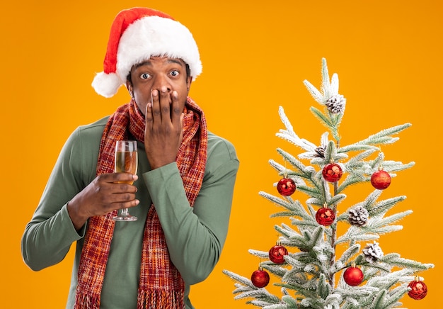 Afro-Amerikaanse man in kerstmuts en sjaal rond nek houden glas champagne kijken camera geschokt bedekking mond met hand naast kerstboom over oranje achtergrond