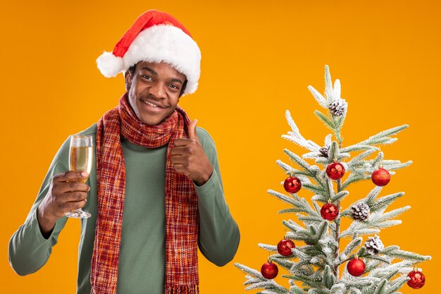 Afro-Amerikaanse man in kerstmuts en sjaal rond nek met glas champagne glimlachend vrolijk weergegeven: duimen naast kerstboom over oranje achtergrond