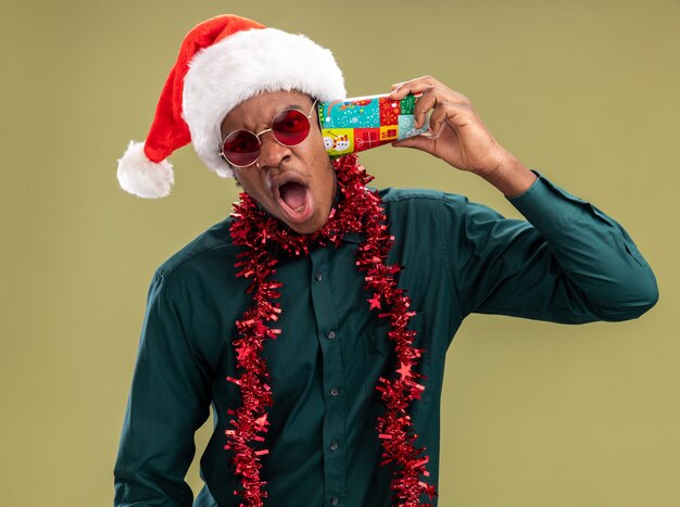 Afro-Amerikaanse man in kerstmuts met slinger dragen van een zonnebril kleurrijke papieren beker houden over zijn oor schreeuwen met geïrriteerde uitdrukking staande over groene achtergrond