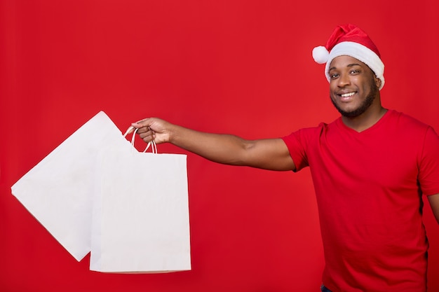 Afro-amerikaanse man lacht in een kerstmuts en houdt witte papieren zakken op een rode achtergrond