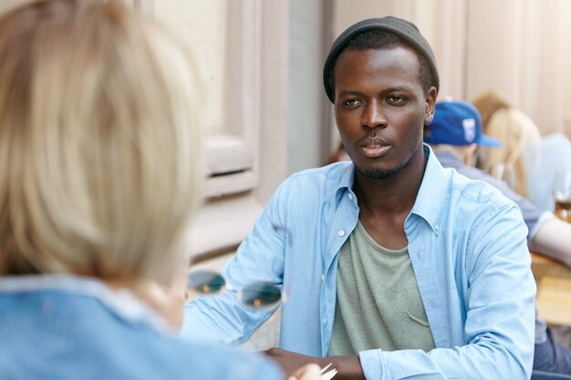 Afro-Amerikaanse man met donkere huid gekleed in shirt en zwarte hoed zitten voor zijn vriendin, gesprek met elkaar, bespreken van nieuws. Zakelijke partners bijeen in café