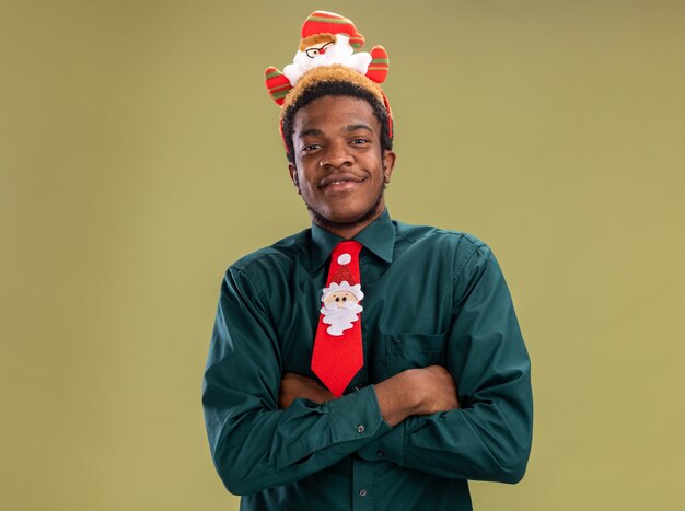 Gratis foto afro-amerikaanse man met grappige santa rand en rode stropdas kijken camera blij en positief glimlachen staande over groene achtergrond