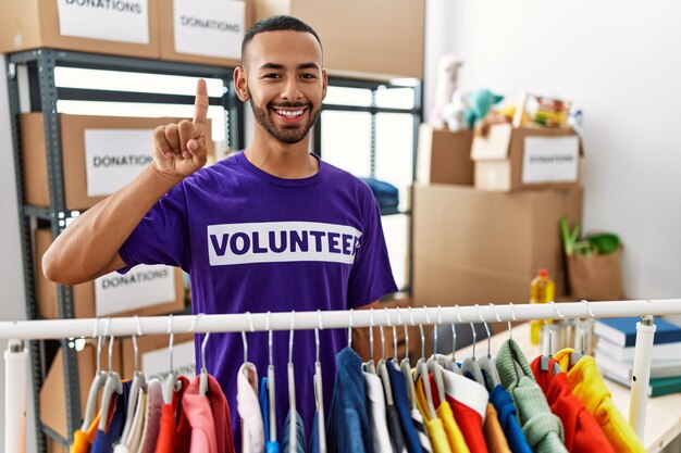 Afro-amerikaanse man met vrijwilligerst-shirt bij donatiestand die toont en omhoog wijst met vinger nummer één terwijl hij zelfverzekerd en gelukkig glimlacht.