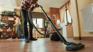 Gratis foto afro-amerikaanse man stofzuigt vloeren in de woonkamer, maakt appartement schoon met vriendin. jonge volwassene die stofzuiger gebruikt en vrouw planken afveegt met allesreiniger. statief opname.