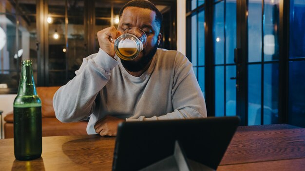 Afro-Amerikaanse man van middelbare leeftijd die bier drinkt met plezier happy night party-evenement online viering via videogesprek in de woonkamer thuis.