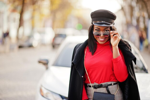 Afro-Amerikaanse mode meisje in jas krantenverkoper pet en zonnebril poseerde op straat tegen witte zakenauto