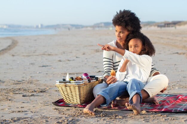 Afro-Amerikaanse moeder en dochter op picknick op het strand. Vrouwen in vrijetijdskleding zittend op deken, broodjes eten, kijken naar water. Familie, ontspanning, natuurconcept