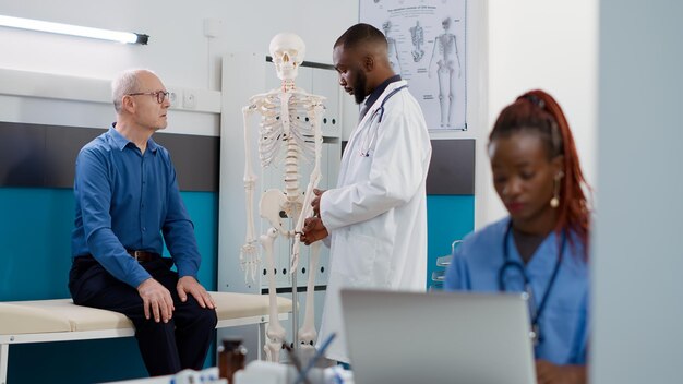Afro-Amerikaanse osteopaat wijzend op menselijk skelet om botziekte en diagnose uit te leggen aan senior patiënt bij controlebezoek. Anatomiespecialist die ruggenmerg toont bij osteopathieonderzoek.