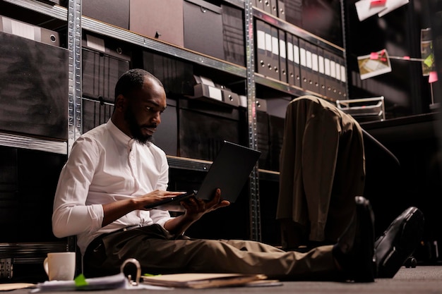 Gratis foto afro-amerikaanse politieagent zittend op de vloer en met behulp van laptop. criminoloog die een misdaaddossier bestudeert en 's nachts informatie op een draagbare computer in een donker kantoor controleert