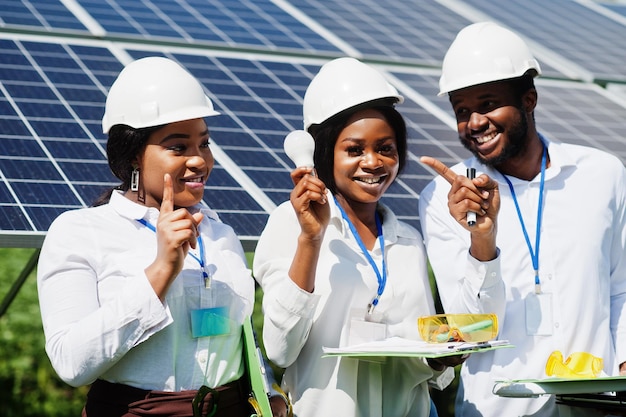 Gratis foto afro-amerikaanse technicus controleert het onderhoud van de zonnepanelen groep van drie zwarte ingenieurs ontmoeten elkaar op zonnestation hold led bulb