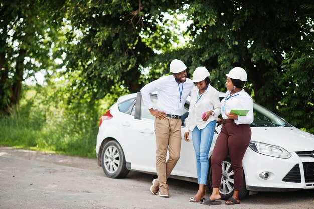 Afro-Amerikaanse technicus in witte helmen in de buurt van auto Groep van drie zwarte ingenieurs die samenkomen