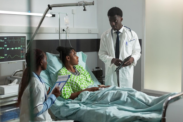 Afro-amerikaanse therapeut arts die zieke vrouw met tablet controleert met medische expertise die de behandeling van de gezondheidszorg bespreekt. patiënt rust in bed tijdens klinische consultatie in ziekenhuisafdeling