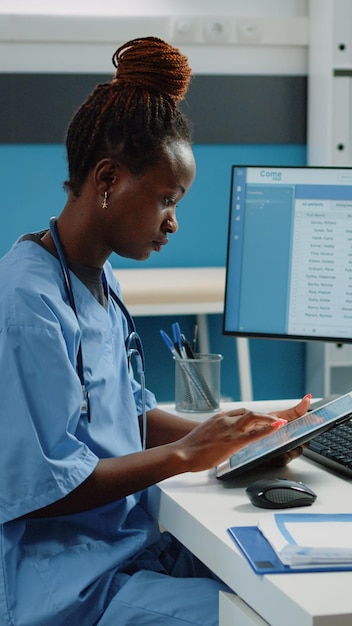 Afro-Amerikaanse verpleegster die digitale tablet gebruikt voor behandeling. Zwarte medische assistent die naar het scherm van het apparaat kijkt terwijl hij een uniform en een stethoscoop draagt in het artsenbureau voor gezondheidszorg.