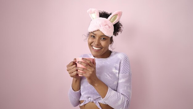 Afro-amerikaanse vrouw die een slaapmasker draagt en een kopje koffie drinkt over een geïsoleerde roze achtergrond