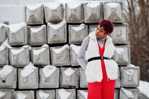 Afro-amerikaanse vrouw in rode broek en wit bontjasje poseerde op winterdag tegen besneeuwde stenen achtergrond