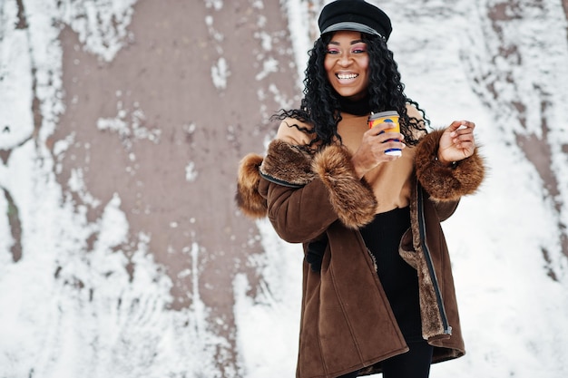 Afro-amerikaanse vrouw in schapenvacht jas en pet poseerde op winterdag tegen besneeuwde achtergrond met kopje koffie