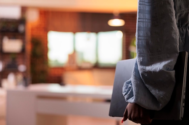 Afro-Amerikaanse vrouw in vrijetijdskleding met laptop in kantoor aan huis met modern interieur en prachtig warm zonsonderganglicht. vrouwelijke ondernemer, vasthouden, draagbare computer, zijaanzicht