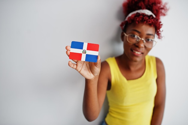 Afro-Amerikaanse vrouw met afro-haar draagt een geel hemd en een bril houdt de vlag van de Dominicaanse Republiek geïsoleerd op een witte achtergrond