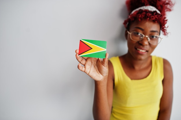 Gratis foto afro-amerikaanse vrouw met afro-haar draagt een geel hemd en een bril houdt de vlag van guyana op een witte achtergrond
