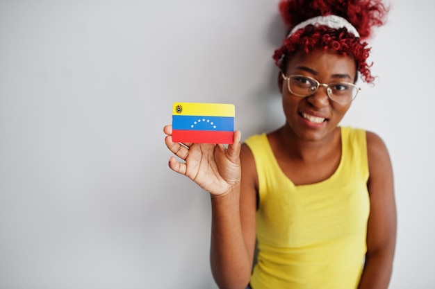 Afro-Amerikaanse vrouw met afro-haar draagt een geel hemd en een bril houdt de vlag van Venezuela vast op een witte achtergrond