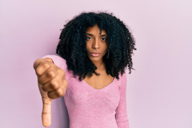 Gratis foto afro-amerikaanse vrouw met afrohaar in een casual roze shirt die er ongelukkig en boos uitziet en afwijzing en negatief toont met duim omlaag gebaar slechte uitdrukking