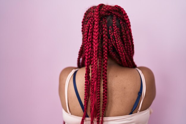 Afro-Amerikaanse vrouw met gevlochten haar die over een roze achtergrond staat en achteruit kijkt met gekruiste armen