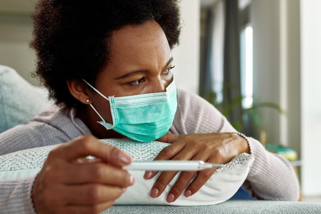 Afro-amerikaanse vrouw met gezichtsmasker voelt zich ontevreden tijdens het gebruik van een thermometer en het controleren van haar temperatuur thuis