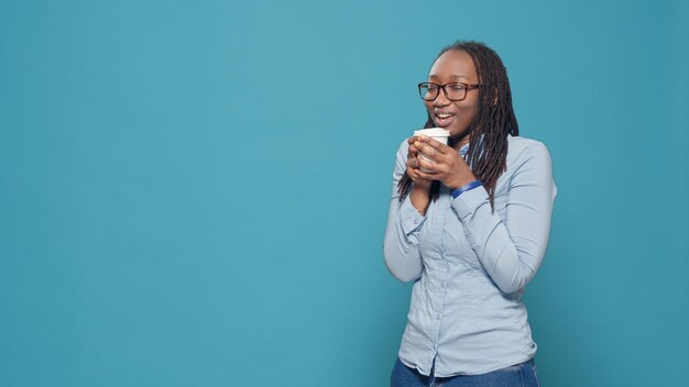 Afro-Amerikaanse vrouw met kopje koffie op camera, opgewonden en blij over het drinken van drank. Genieten van verfrissing van warme dranken en plezier maken tijdens de pauze op de achtergrond van de studio.